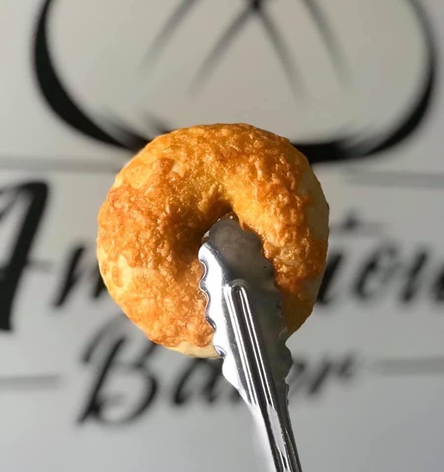 A cheddar-crusted sourdough bagel held on tongs in front of the Ambitious Baker logo